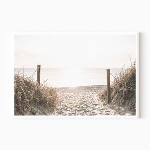 May include: A pathway through tall grasses leads to a sandy beach with a view of the ocean and a bright, hazy sky.
