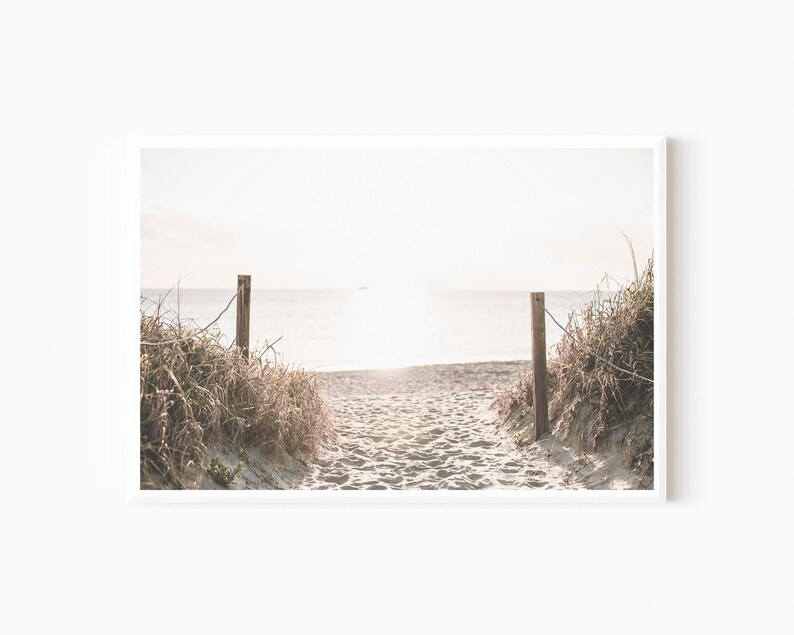 May include: A pathway through tall grasses leads to a sandy beach with a view of the ocean and a bright, hazy sky.