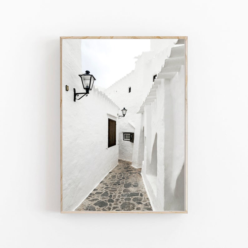 May include: A narrow, white-washed alleyway with a cobblestone floor.  The walls are lined with traditional lanterns and the alleyway leads to a bright, open space.
