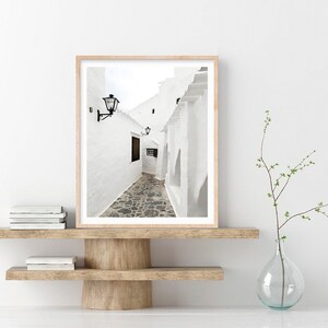 May include: A black and white photograph of a narrow, white-walled alleyway in a European village. The alleyway is lined with buildings and has a cobblestone floor. The photograph is framed in a light wood frame.