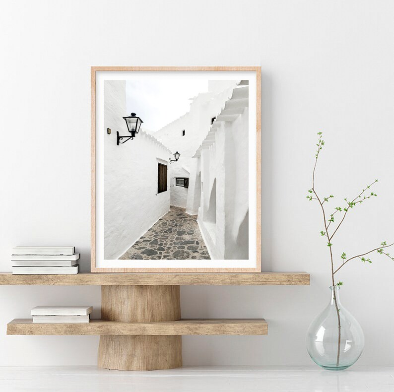 May include: A black and white photograph of a narrow, white-walled alleyway in a European village. The alleyway is lined with buildings and has a cobblestone floor. The photograph is framed in a light wood frame.
