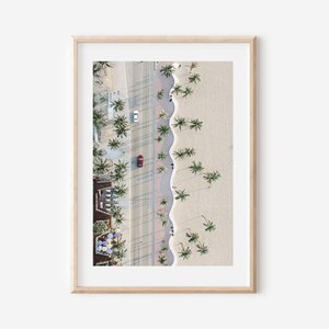 May include: An aerial view of a beach with palm trees lining the shore. A red car is driving down the road next to the beach. The beach is sandy and the water is blue.