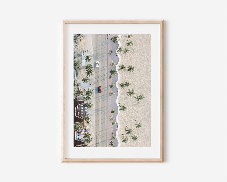 May include: An aerial view of a beach with palm trees lining the shore. A red car is driving down the road next to the beach. The beach is sandy and the water is blue.