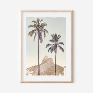 May include: A framed photograph of two palm trees in front of a rocky mountain. The sky is a light blue and the mountain is a light brown. The photograph is in a light wood frame.