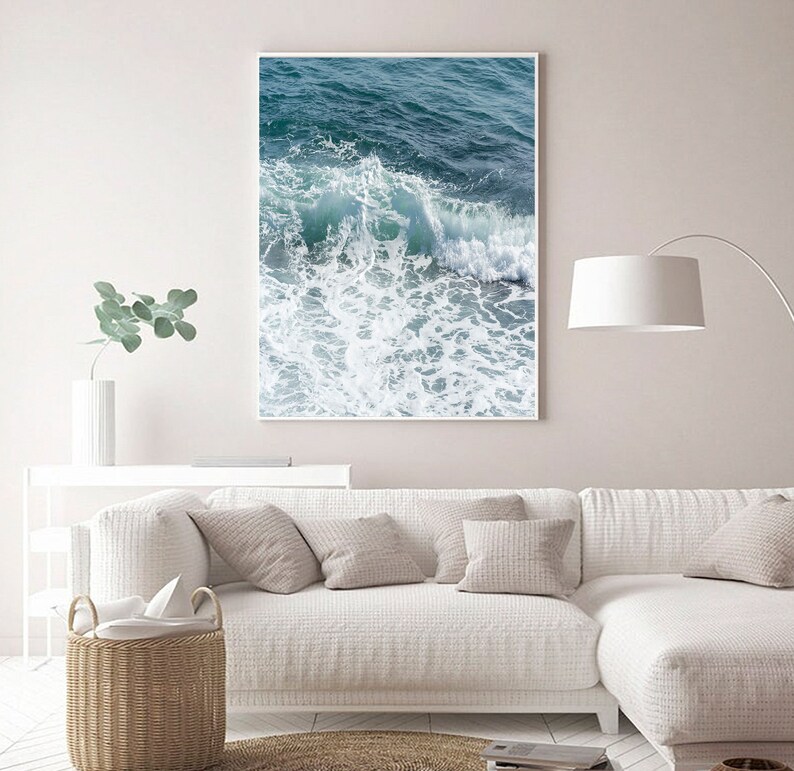 May include: A framed print depicting a breaking ocean wave. The artwork shows turquoise and deep blue water with white foam. The print hangs above a white sofa with neutral cushions, creating a calm living room setting.