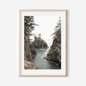 May include: Framed landscape print featuring a coastal scene. The artwork depicts rocky cliffs with evergreen trees, framing a view of the ocean and small islands. The color palette is muted, with shades of gray, green, and blue.