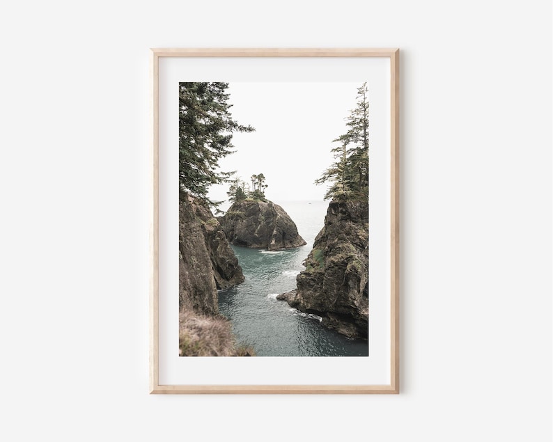 May include: Framed landscape print featuring a coastal scene. The artwork depicts rocky cliffs with evergreen trees, framing a view of the ocean and small islands. The color palette is muted, with shades of gray, green, and blue.