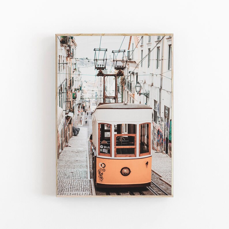 May include: A yellow and white vintage tram travels up a cobblestone street in a European city. The tram has a sign that says "Wi-Fi" on the side. The street is lined with buildings on both sides.