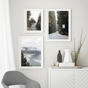 May include: Three framed landscape prints. One shows a coastal view with rocky cliffs and water. Another depicts a road through a forest. The third displays a misty beach scene. The frames are white, and the overall aesthetic is minimalist.