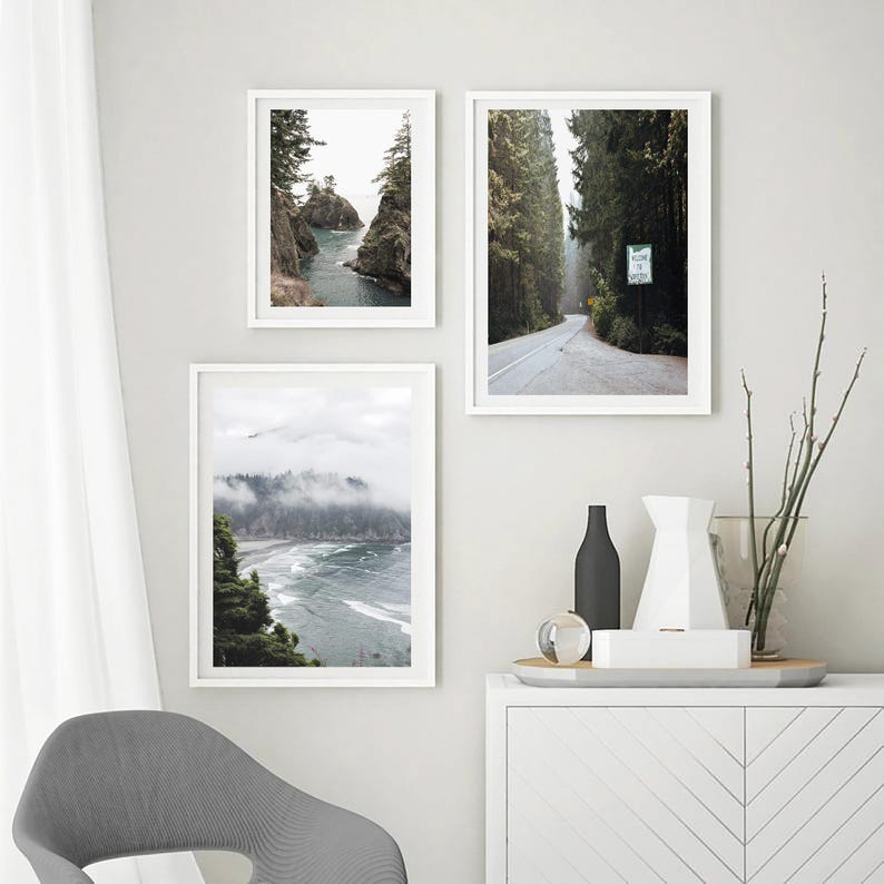 May include: Three framed landscape prints. One shows a coastal view with rocky cliffs and water. Another depicts a road through a forest. The third displays a misty beach scene. The frames are white, and the overall aesthetic is minimalist.