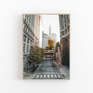 May include: A view down a city street with tall buildings on either side. The street is empty and there are trees lining the sides. The sky is a light blue and there are clouds in the distance.