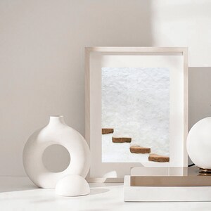 Puede incluir: Un jarr&oacute;n de cer&aacute;mica blanco con una abertura circular en el medio, una esfera de cer&aacute;mica blanca y una fotograf&iacute;a enmarcada de una pared blanca con un conjunto de escalones marrones. La fotograf&iacute;a est&aacute; sobre una superficie dorada y blanca.