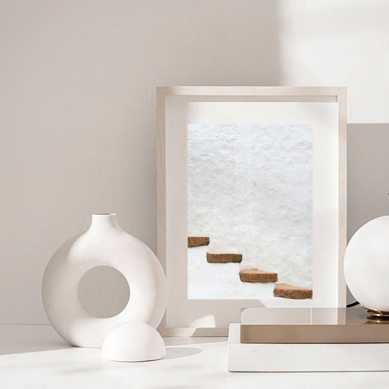 Puede incluir: Un jarr&oacute;n de cer&aacute;mica blanco con una abertura circular en el medio, una esfera de cer&aacute;mica blanca y una fotograf&iacute;a enmarcada de una pared blanca con un conjunto de escalones marrones. La fotograf&iacute;a est&aacute; sobre una superficie dorada y blanca.