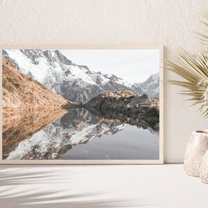 Puede incluir: Una fotograf&iacute;a enmarcada de un lago de monta&ntilde;a con picos cubiertos de nieve que se reflejan en el agua. El lago est&aacute; rodeado de colinas verdes y una peque&ntilde;a caba&ntilde;a de madera es visible en el lado derecho de la imagen.