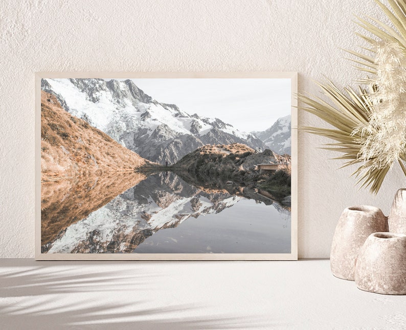 Puede incluir: Una fotograf&iacute;a enmarcada de un lago de monta&ntilde;a con picos cubiertos de nieve que se reflejan en el agua. El lago est&aacute; rodeado de colinas verdes y una peque&ntilde;a caba&ntilde;a de madera es visible en el lado derecho de la imagen.