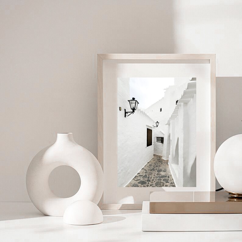 May include: A framed photograph of a white alleyway with a cobblestone path and black lanterns on the walls. The photo is displayed on a white surface with a white vase and a white sphere.