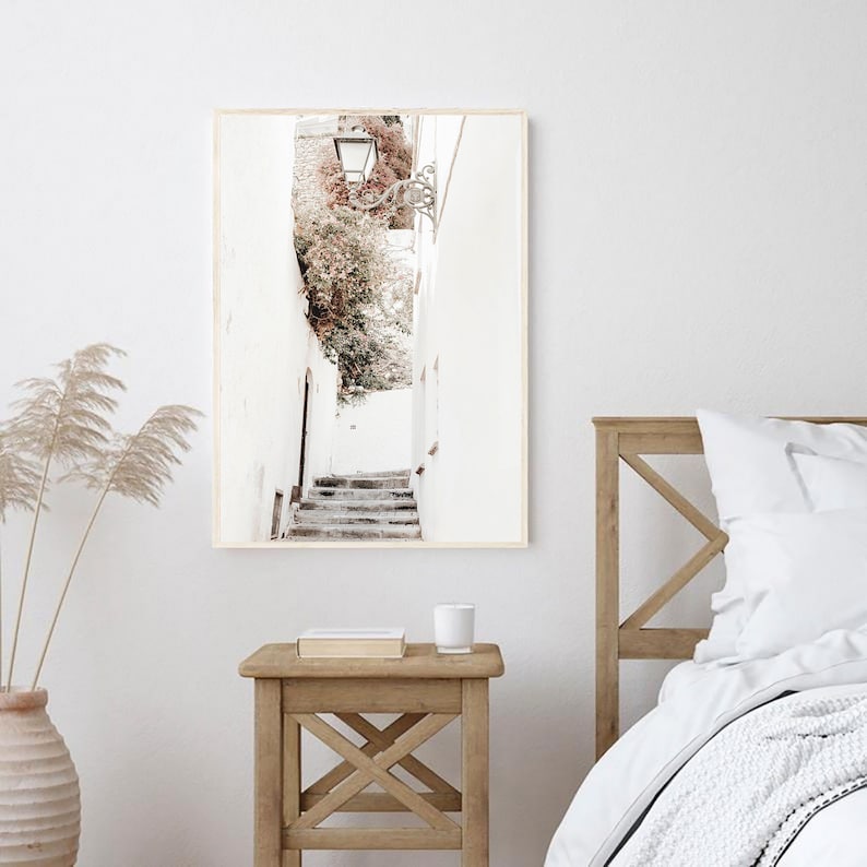 May include: A framed photograph of a white alleyway with a stone staircase leading up to a building with a leafy green plant growing on the side. The alleyway is lit by a street lamp.