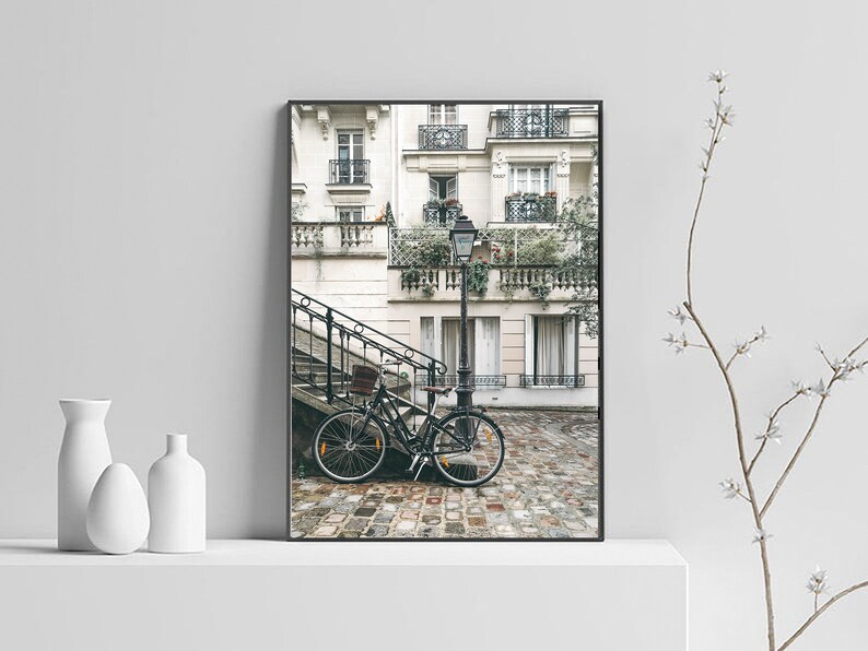 May include: A black bicycle is parked on a cobblestone street in front of a white building with a black wrought iron railing. The building has multiple windows and balconies with plants growing on them. A street lamp is in the background.
