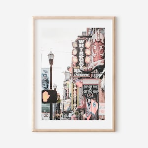 May include: A vintage-style photograph of a street scene in Nashville, Tennessee. The image features a row of brick buildings with neon signs, including a sign for a "Whiskey Saloon" with a large red cowboy boot. The signs are lit up and create a vibrant atmosphere.