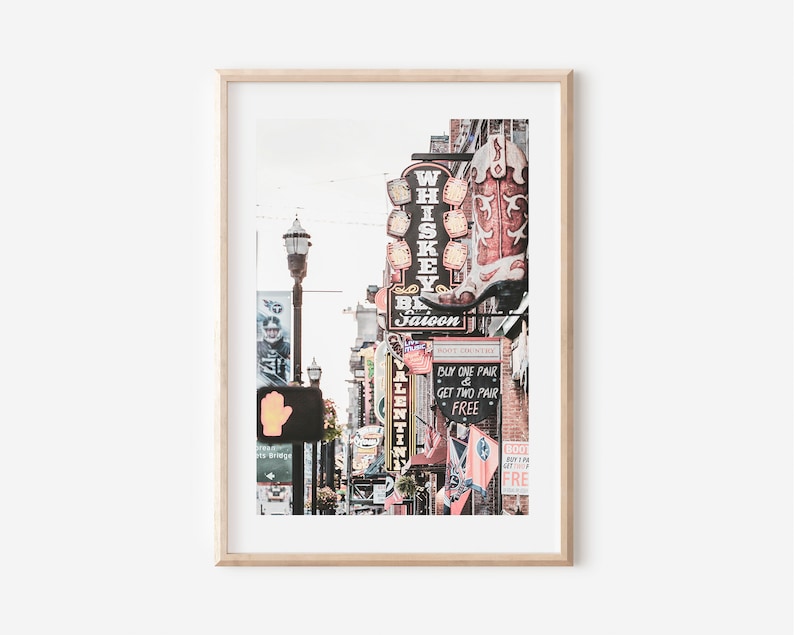 May include: A vintage-style photograph of a street scene in Nashville, Tennessee. The image features a row of brick buildings with neon signs, including a sign for a "Whiskey Saloon" with a large red cowboy boot. The signs are lit up and create a vibrant atmosphere.