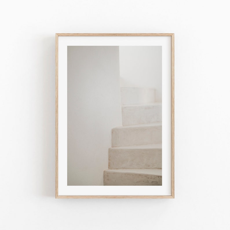May include: A framed print depicting a minimalist architectural photograph. The image shows a section of a white staircase against a white wall, creating a sense of depth and simplicity. The frame is a light wood colour.