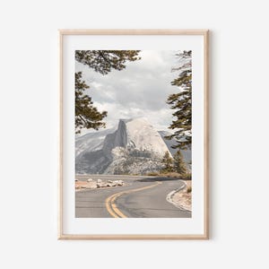 May include: Framed landscape print featuring a winding road leading towards a large mountain. The image is framed by trees and a cloudy sky. The road has yellow lines and is surrounded by rocks and vegetation. The overall color palette is muted with natural tones.