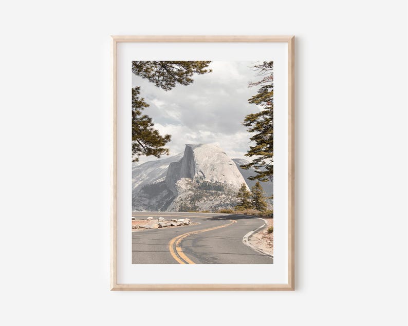 May include: Framed landscape print featuring a winding road leading towards a large mountain. The image is framed by trees and a cloudy sky. The road has yellow lines and is surrounded by rocks and vegetation. The overall color palette is muted with natural tones.
