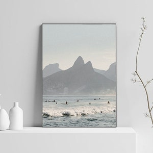 May include: A photograph of a beach scene with a mountain range in the background. The water is blue and there are waves crashing on the shore. There are people surfing in the water.