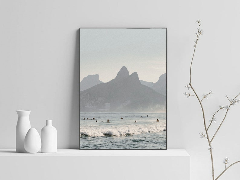 May include: A photograph of a beach scene with a mountain range in the background. The water is blue and there are waves crashing on the shore. There are people surfing in the water.