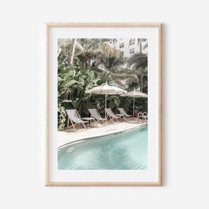 May include: A framed print of a tropical pool scene with lounge chairs and umbrellas. The pool is surrounded by lush greenery and palm trees.