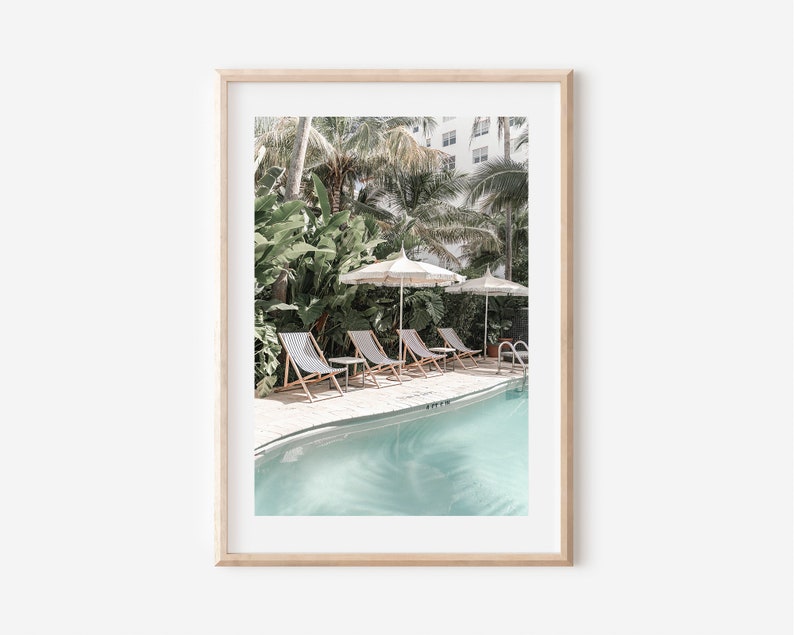 May include: A framed print of a tropical pool scene with lounge chairs and umbrellas. The pool is surrounded by lush greenery and palm trees.