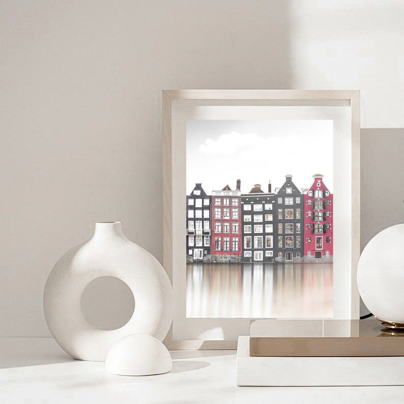 May include: A framed print of a row of colorful buildings reflected in a canal. The buildings are a mix of red, black, and white. The print is in a white frame and is sitting on a white surface.