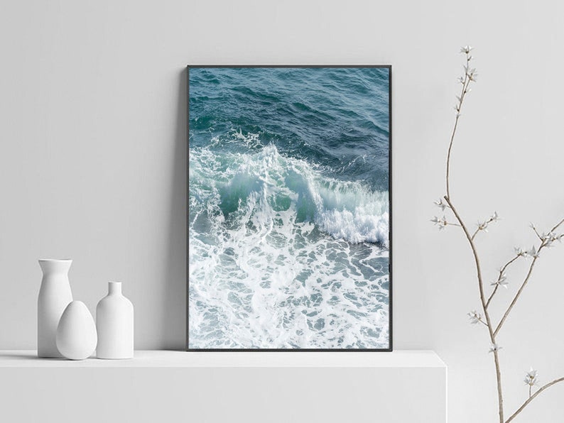 May include: A framed print showing a close-up of ocean waves. The image displays the turquoise and deep blue colours of the water, with white foamy crests. The artwork is displayed on a white shelf, with white vases and a decorative branch.