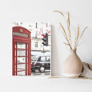 May include: A red telephone booth with the word "TELEPHONE" in gold lettering on the front. The booth is on a street in London, England, with a black taxi cab in the background.