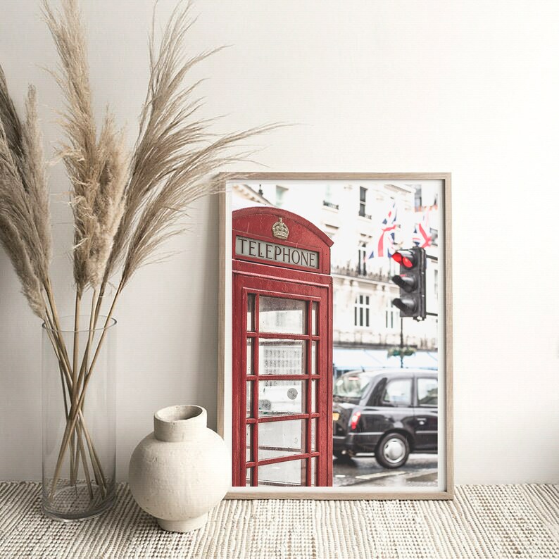 May include: A red telephone booth with the word "TELEPHONE" on the front, located on a street in London, England. The booth is in front of a black taxi cab and a traffic light.