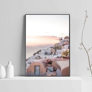 May include: A photograph of a white and pink village with a blue ocean in the background. The village is built on a cliffside and has a windmill on top of a hill. The sky is a light blue with pink and orange clouds.