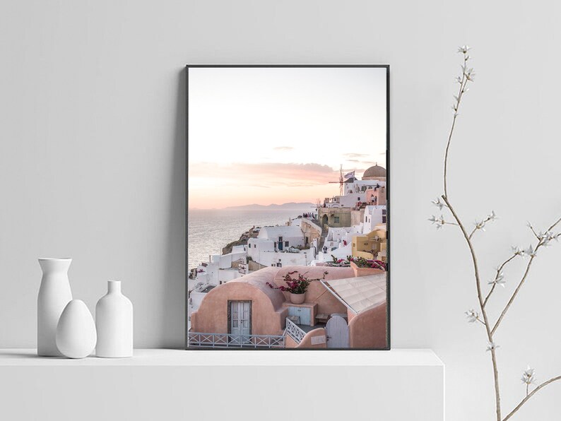 May include: A photograph of a white and pink village with a blue ocean in the background. The village is built on a cliffside and has a windmill on top of a hill. The sky is a light blue with pink and orange clouds.
