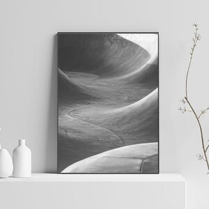 May include: Black and white photograph of a concrete skate park with a series of curved ramps. The photo is framed in a black frame and is displayed on a white shelf.
