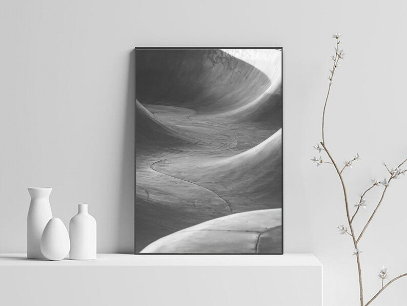 May include: Black and white photograph of a concrete skate park with a series of curved ramps. The photo is framed in a black frame and is displayed on a white shelf.