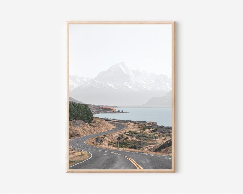 May include: A winding road leads through a mountain landscape with a lake in the distance. The mountains are snow-capped and the sky is a light blue.