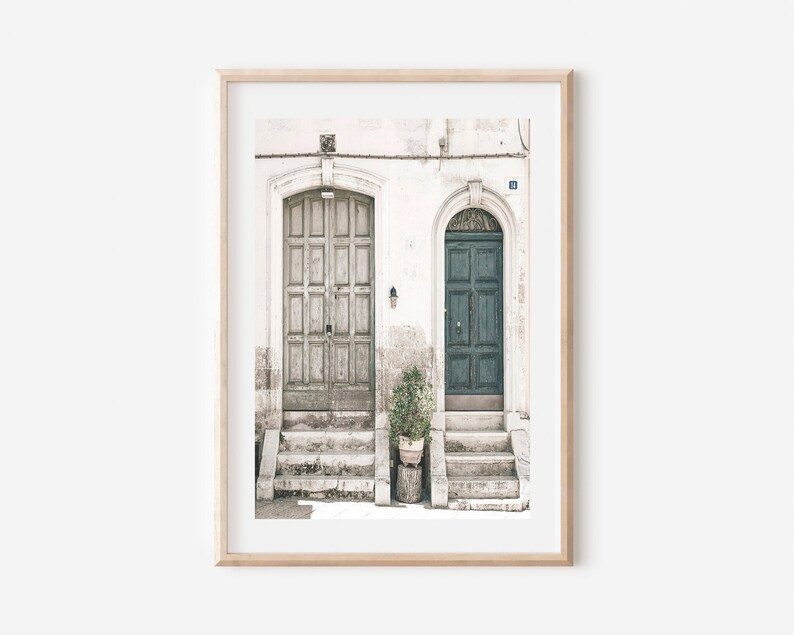May include: Two doors, one painted white and the other a deep green, are set into a white building with a potted plant in front of the green door. The building has a weathered look and the doors are both slightly ajar.