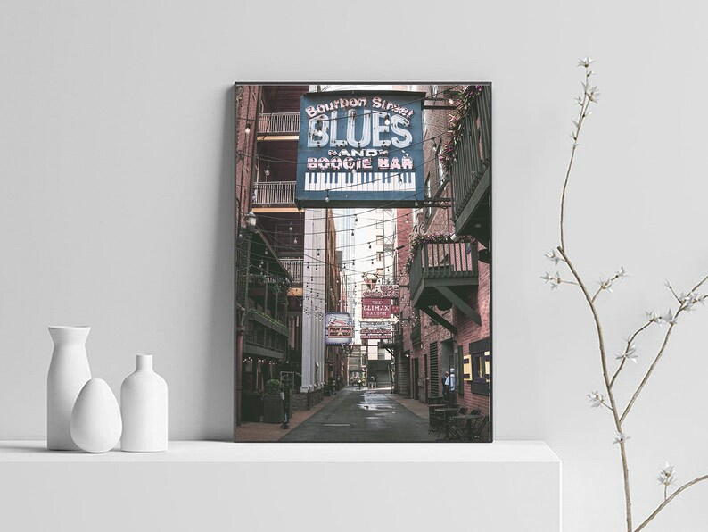 Puede incluir: Un letrero vintage para "Bourbon Street Blues Piano and Boogie Bar" cuelga sobre un callej&oacute;n estrecho y adoquinado. El letrero es azul con letras blancas y presenta un gr&aacute;fico de teclado de piano. El callej&oacute;n est&aacute; iluminado con luces de cadena y tiene un ambiente r&uacute;stico y urbano.