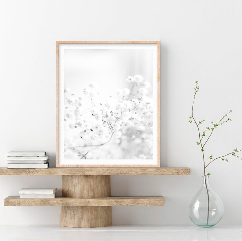 Pode incluir: Uma fotografia a preto e branco de um delicado cacho de flores, enquadrada numa moldura de madeira clara e exposta numa prateleira de madeira com um vaso de vidro transparente e um ramo com folhas verdes.
