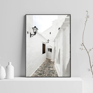 May include: A black and white photograph of a narrow, white-walled alleyway with a stone floor. The alleyway is lined with buildings on both sides, and there is a street lamp hanging from the wall on the left side of the image.