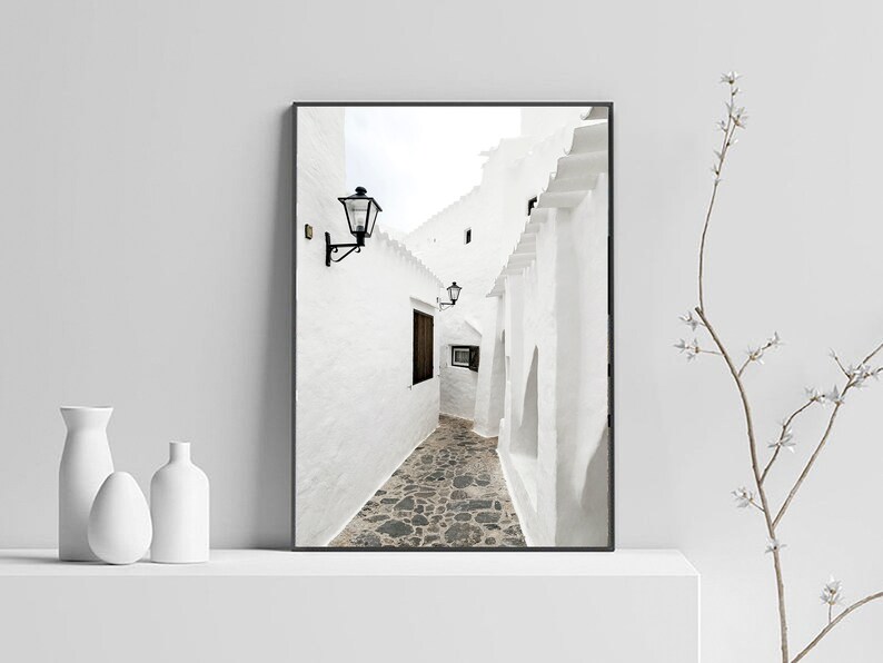 May include: A black and white photograph of a narrow, white-walled alleyway with a stone floor. The alleyway is lined with buildings on both sides, and there is a street lamp hanging from the wall on the left side of the image.