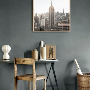 May include: A framed photograph of the New York City skyline with the Empire State Building in the center. The photo is in a light wood frame and is hanging on a gray wall.