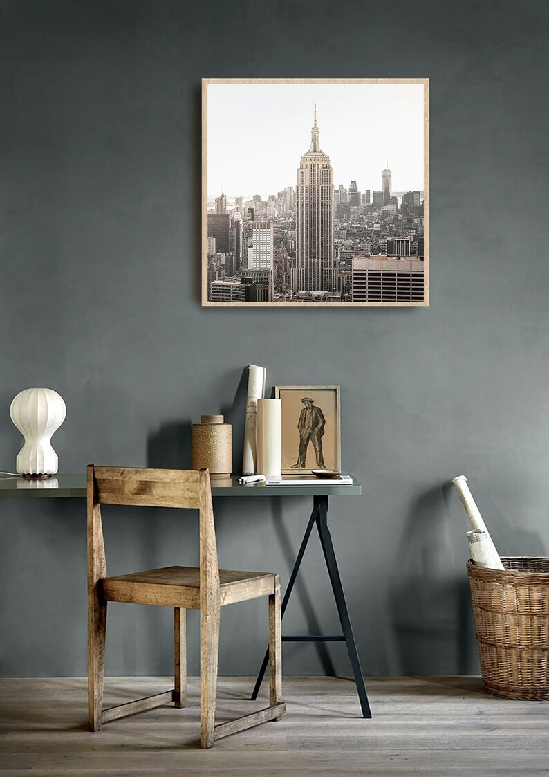 May include: A framed photograph of the New York City skyline with the Empire State Building in the center. The photo is in a light wood frame and is hanging on a gray wall.
