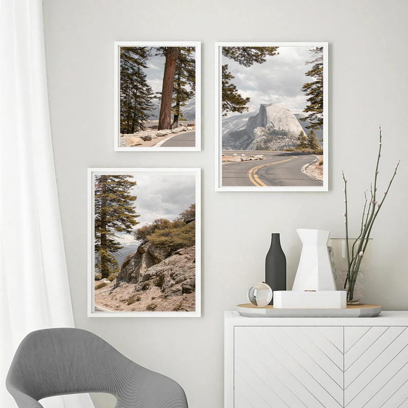 May include: Three framed landscape prints featuring scenic views of a mountain road and towering trees. The prints are in white frames and are displayed on a white wall. The images have a neutral color palette with a focus on natural elements.