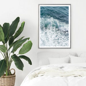 May include: A framed print of ocean waves in teal and white hangs on a white wall. The artwork is above a bed with white linens and pillows. A large green plant in a woven basket is in the foreground.