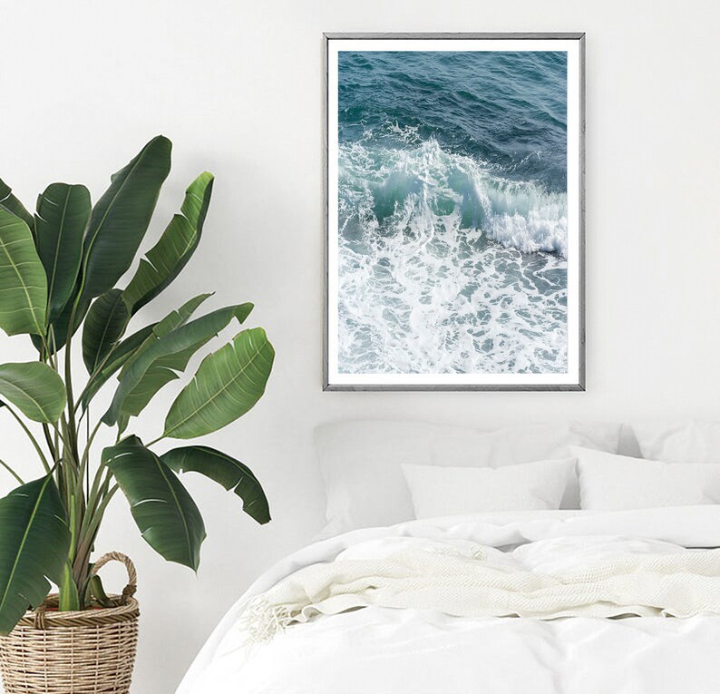 May include: A framed print of ocean waves in teal and white hangs on a white wall. The artwork is above a bed with white linens and pillows. A large green plant in a woven basket is in the foreground.